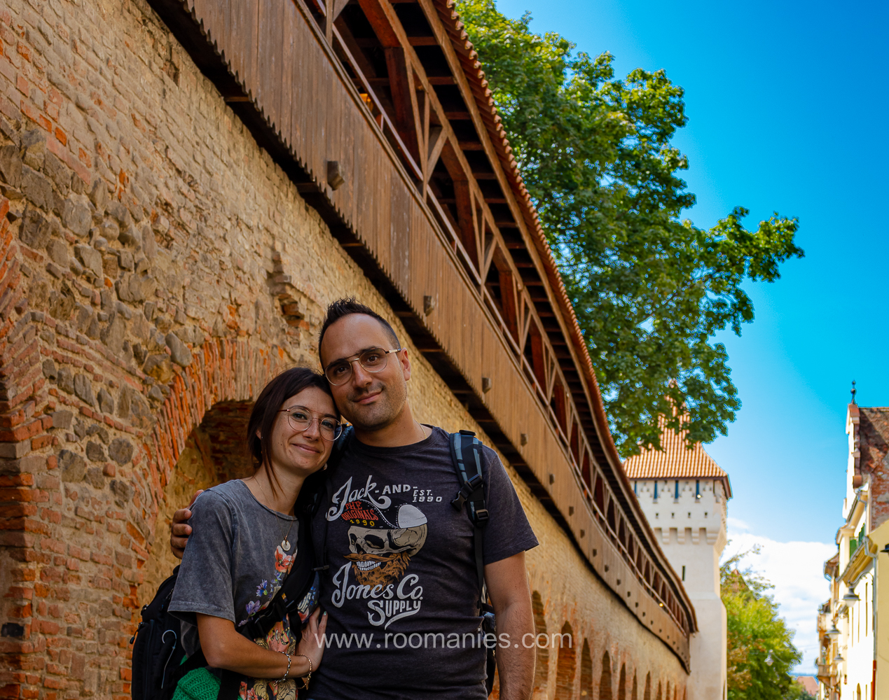 En premier plan Emelin et Laurant regardant vers la caméra et en souriant. En arrière-plan la rue Cetații de Sibiu, avec ses murs en pierre rouge et l’une des tours de la rue. 