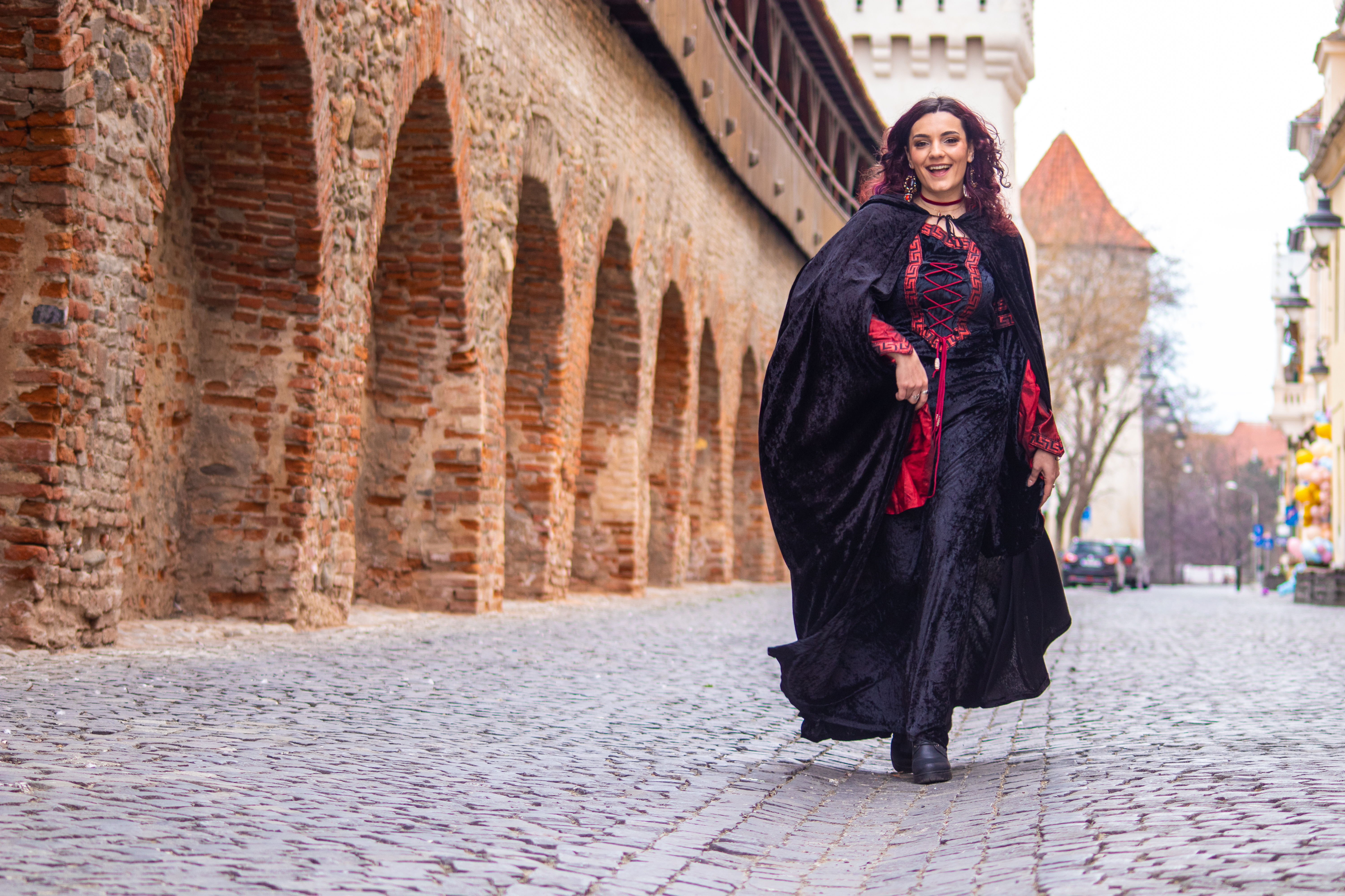 Jeune femme vêtue en tenue médiévale au premier plan. En arrière-plan les murs rougeatres et l’ une des tours médiévales de Sibiu.
