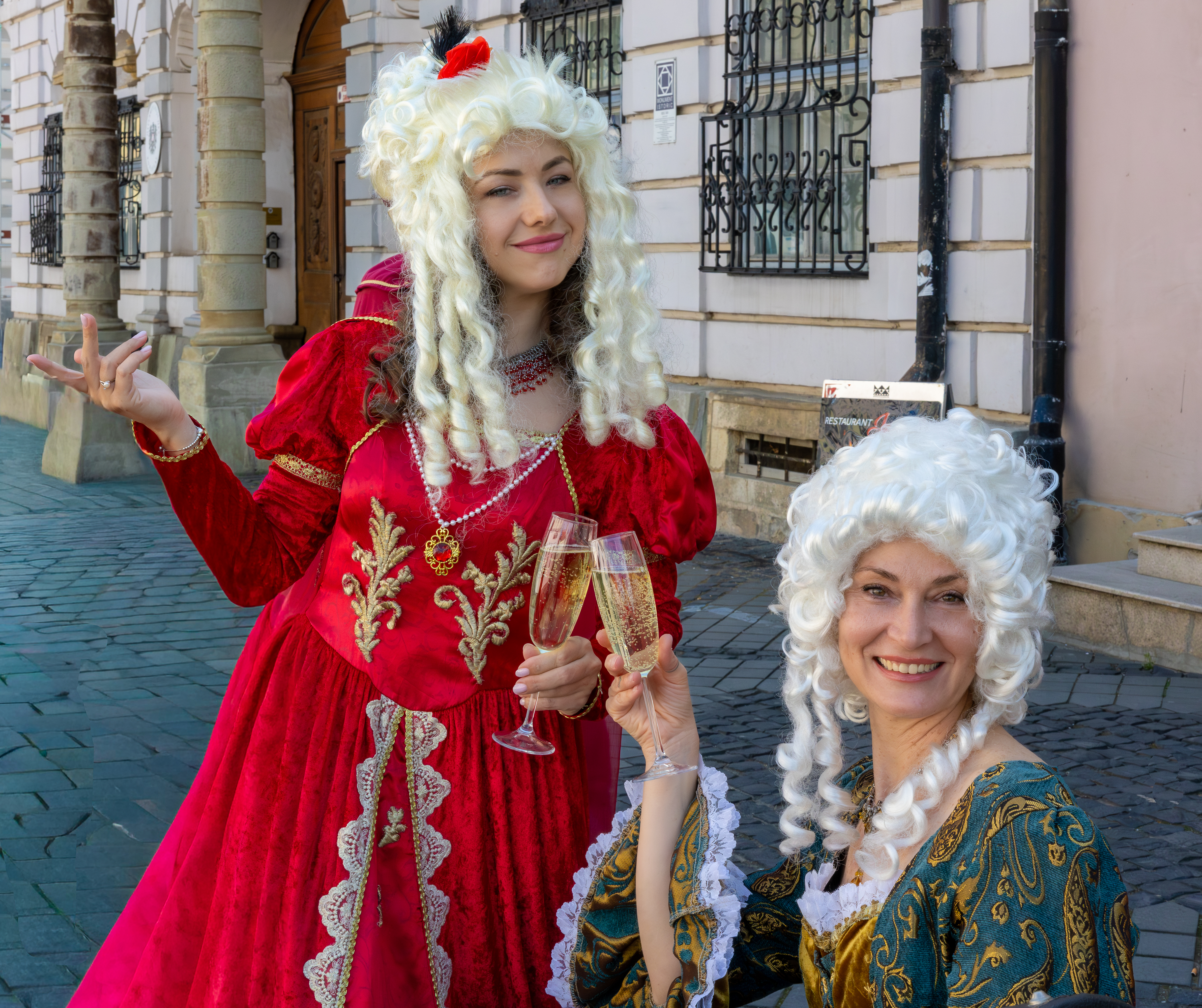 Deux femmes vêtues de tenues médiévales – deux marquises au premier plan. Elles tiennent un verre de champagne a la main, et affichent un large sourire vers la caméra.