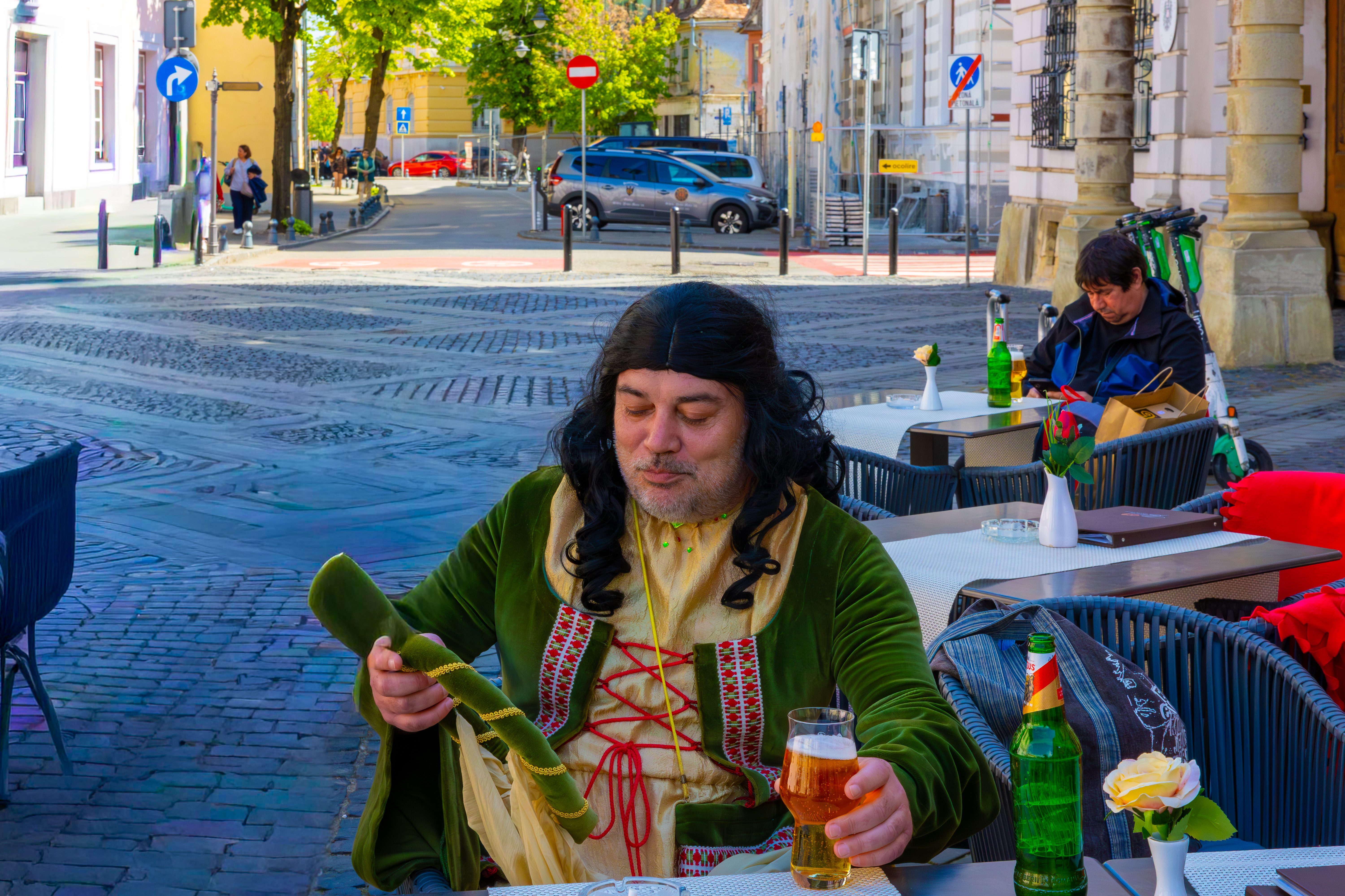 Un homme, vêtu d’une tenue médiévale de femme. Il porte même une perruque. Il est assis sur une terrasse du centre-ville de Sibiu, une bière à la main.