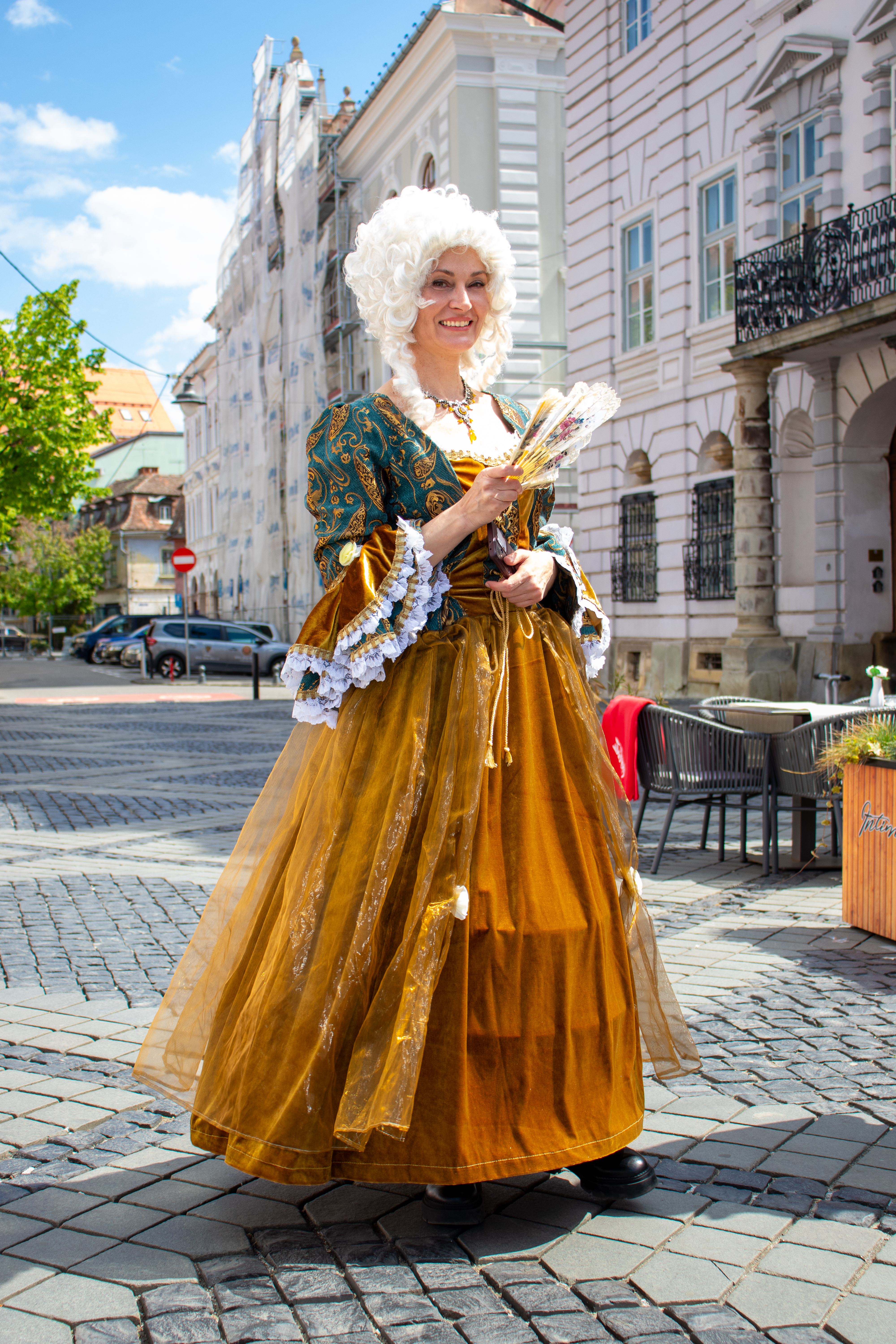 Une femme vêtue en tenue médiévale – une marquise au premier plan. Elles se trouve sur une rue de Sibiu et tient un verre éventail à la main, et affichent un sourire vers la caméra.