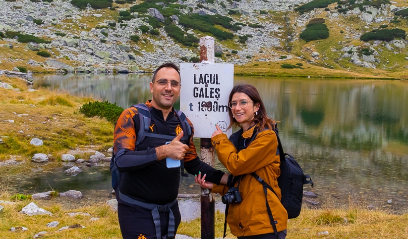 Émeline et Laurent juste à côté du panneau indiquant l’altitude du lac Gales du Retezat, Roumanie, avec en arrière-plan une petite partie du lac Gales et des rochers du Retezat.  