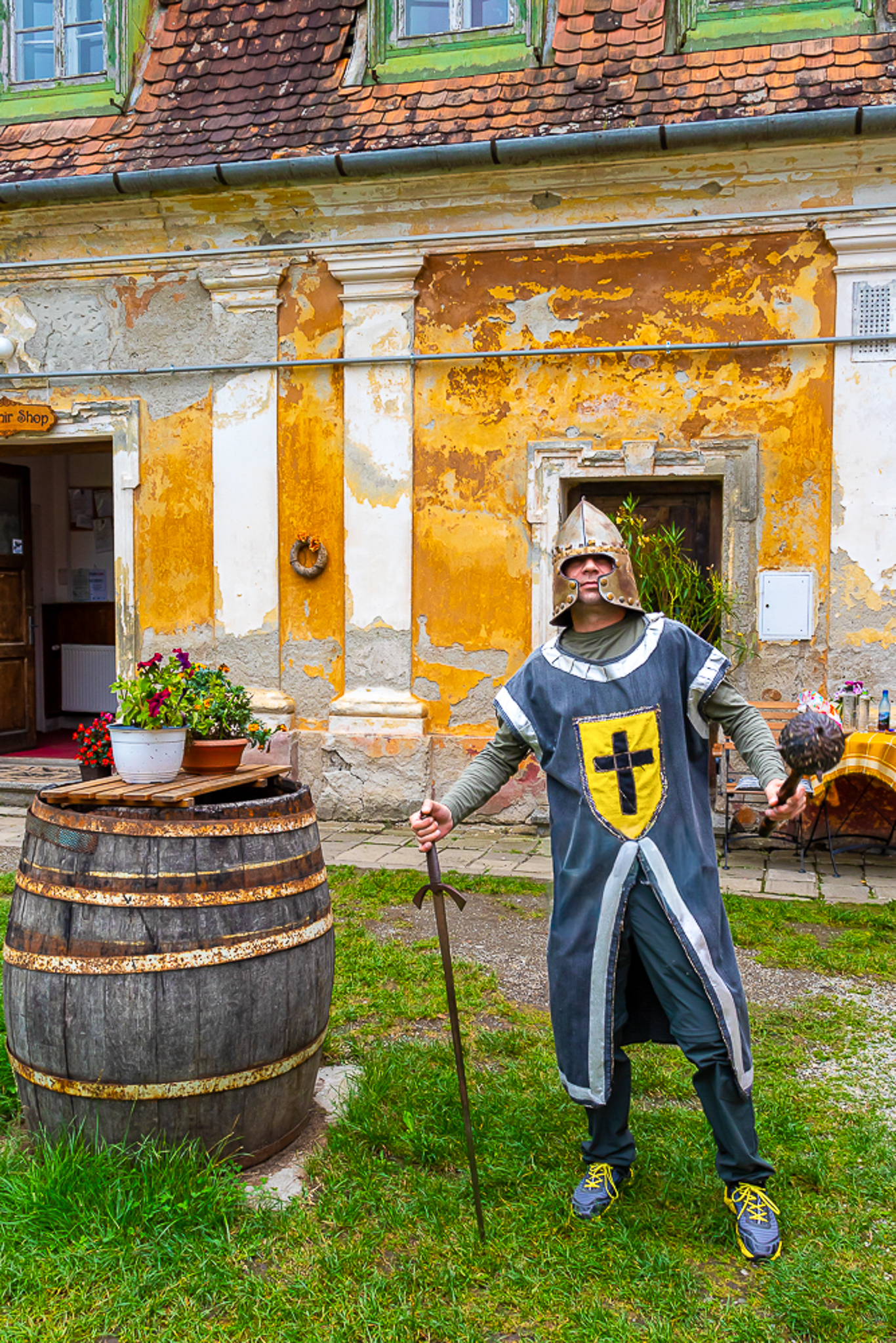 Un homme en armure médiévale tenant une épée et une massue, debout à côté d'un tonneau en bois, devant un bâtiment ancien aux murs peints en jaune et blanc.