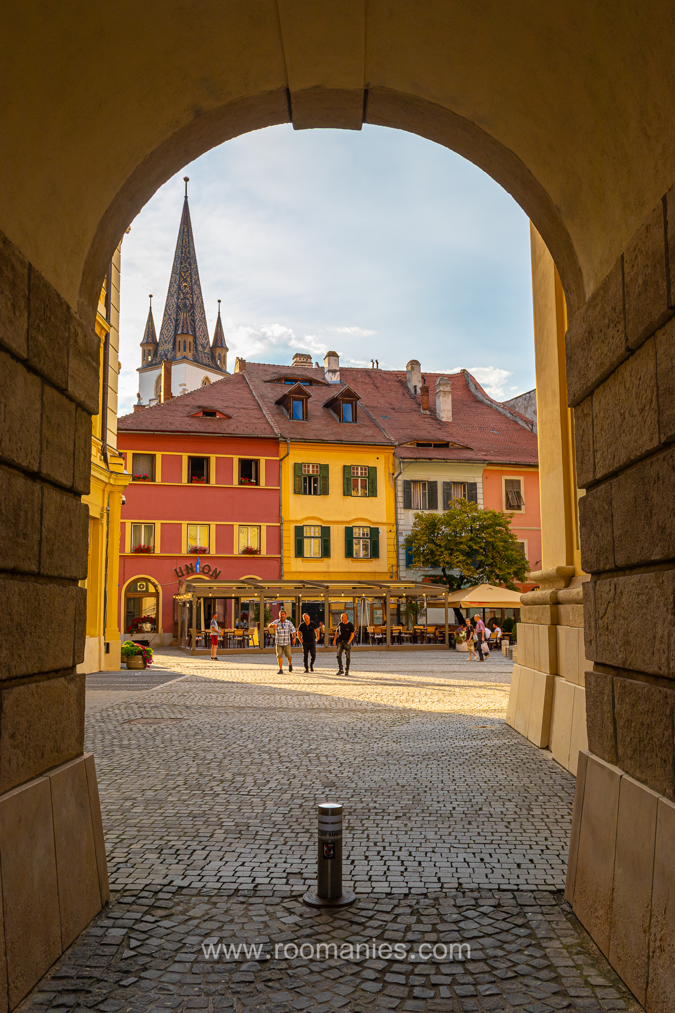 Au premier plan, l’une des arches emblématiques de Sibiu, un passage voûté reliant la Grande Place et la Petite Place ; en arrière-plan quelques maisons médiévales colorées qui se trouvent sur la Petite Place.