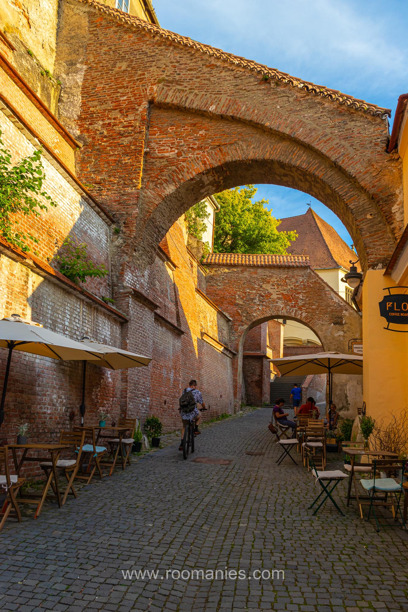 Le Passage des Escaliers de Sibiu, avec au premier plan les tables des restaurants, un homme sur un vélo et la petite ruelle avec ses murs en pierre rouge et ses arches médiévales. En arrière-plan deux humains attablés et la ruelle avec ses murs rouges.