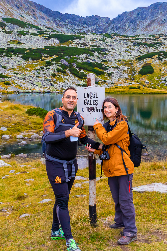 Deux personnes posant devant un panneau indicatif du Lac Gales, à 1900 mètres d'altitude, avec des montagnes et un lac en arrière-plan.