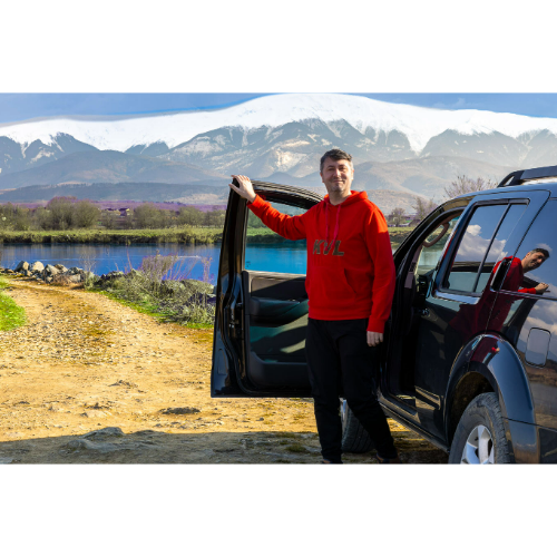 Un jeune homme se tient à côté d'une voiture noire, avec la porte ouverte, devant un paysage de montagne enneigée et un lac.