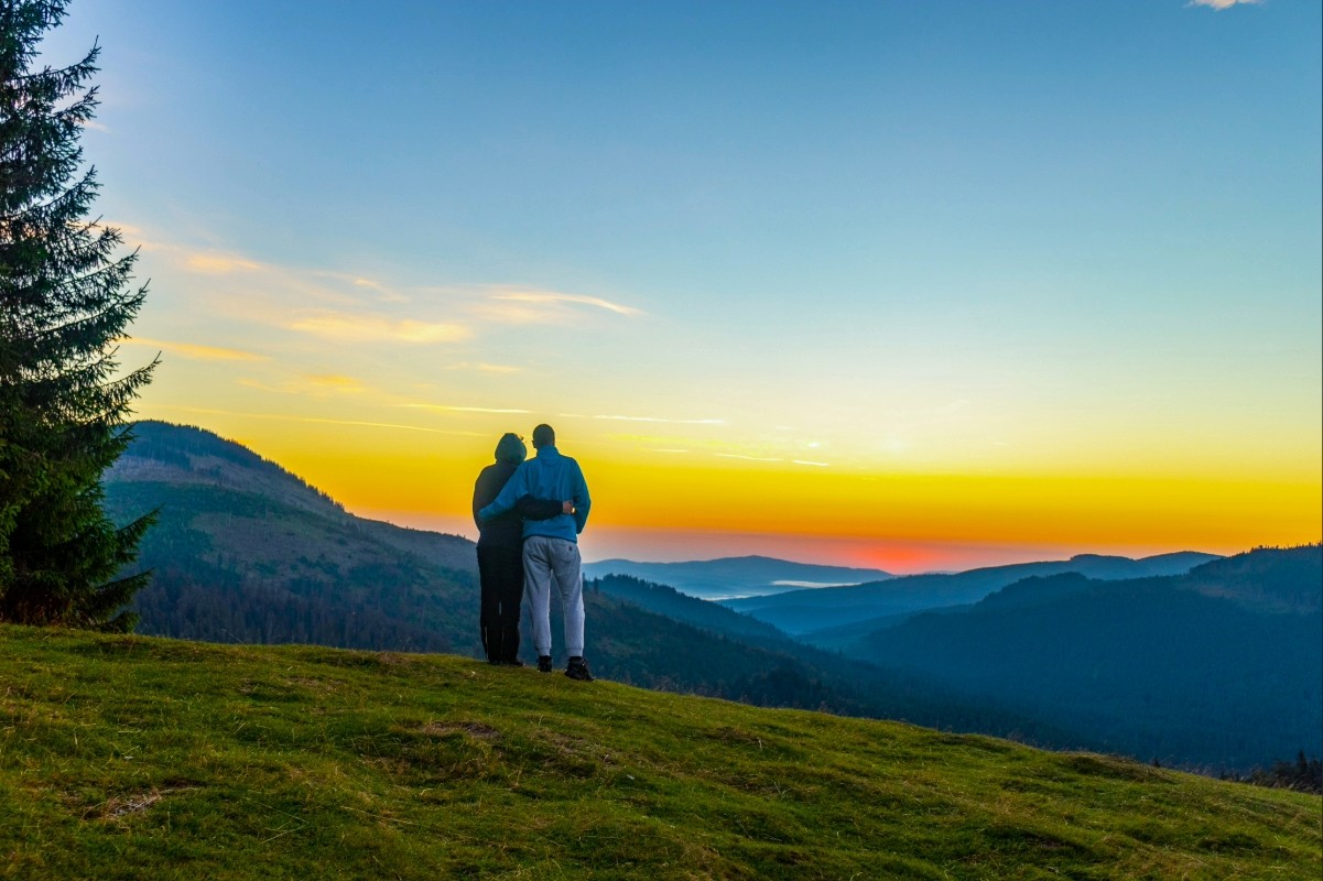 Un couple regardant un coucher de soleil sur les montagnes depuis une colline, entouré de verdure.