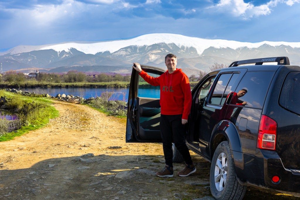 Un homme en sweat à capuche rouge se tient à côté d'un véhicule tout-terrain noir, avec des montagnes enneigées en arrière-plan, près d'un lac et d'un chemin de terre. Le vehicule tout-terrain est
celui utilisé pour les explorations sauvages .