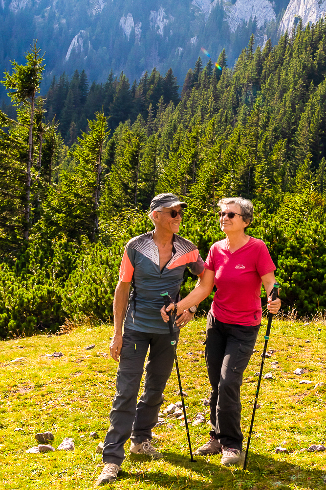 Deux randonneurs souriants sur un sentier montagneux entouré d'arbres et de paysages verdoyants.