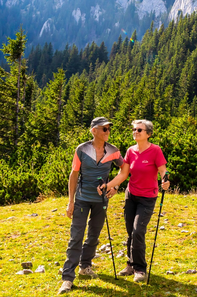 Un couple souriant en randonnée dans les montagnes, portant des vêtements de plein air et tenant des bâtons de randonnée, avec une forêt verdoyante en arrière-plan.