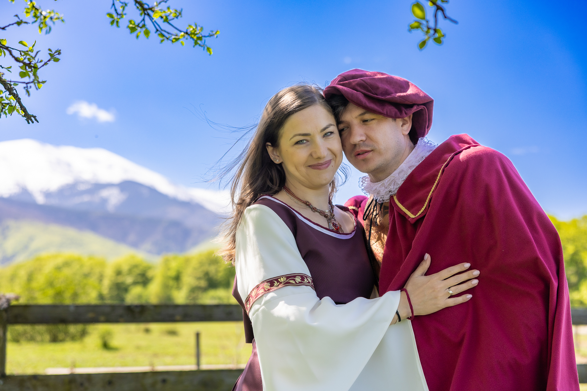 Un couple en costumes médiévaux pose devant un paysage avec une montagne enneigée en arrière-plan, sous un ciel bleu.