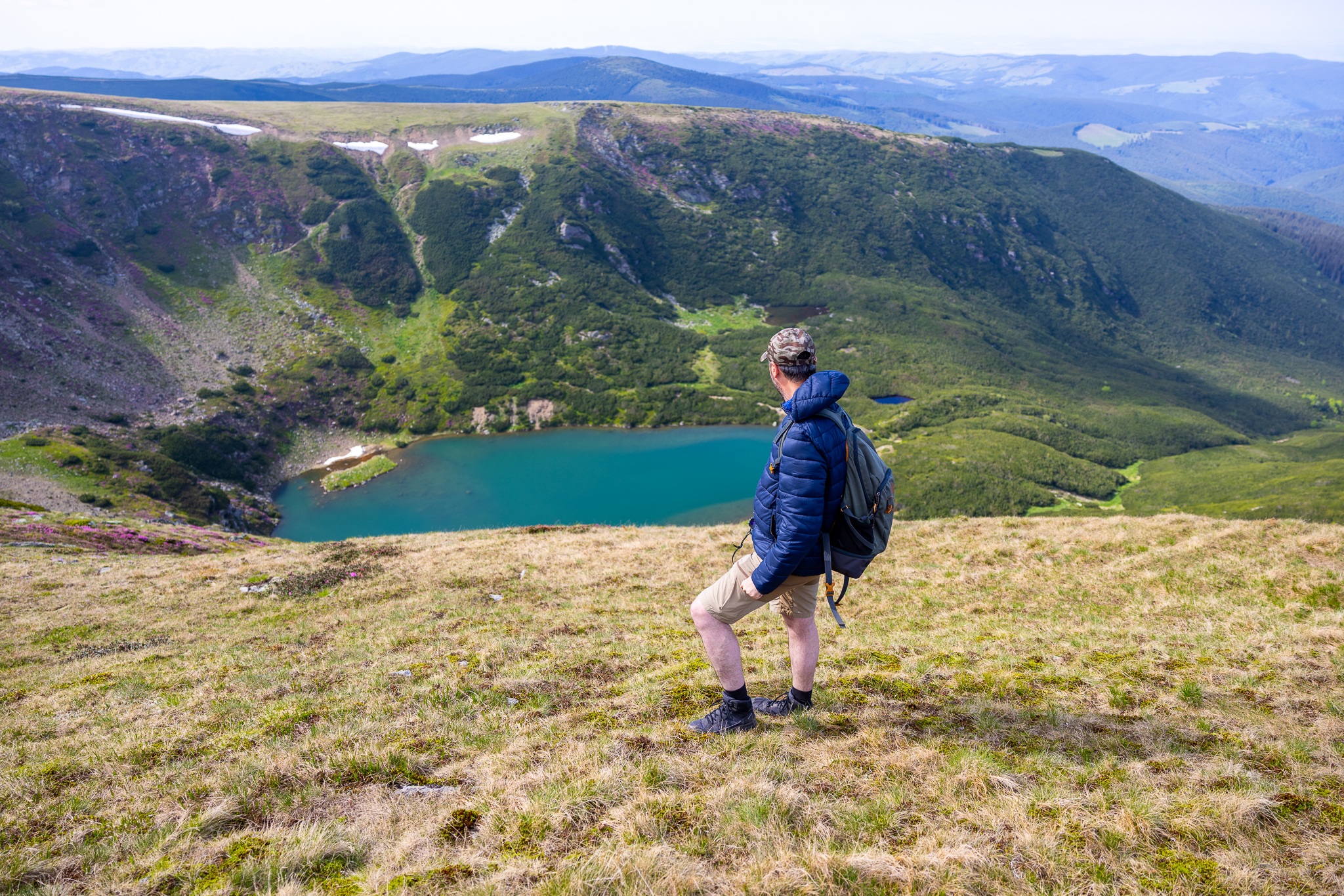 Un randonneur contemplant un lac turquoise dans une vallée verdoyante, entouré de montagnes.